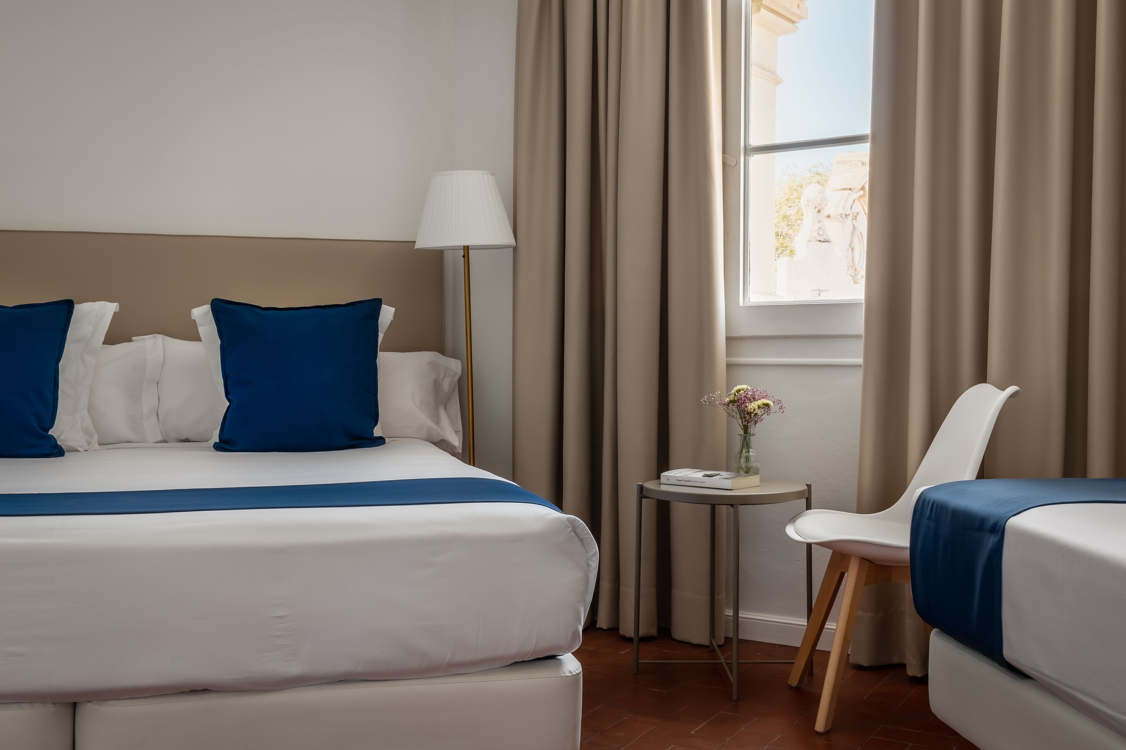 A hotel room with a bed, a chair, a lamp, and a small table with a vase.