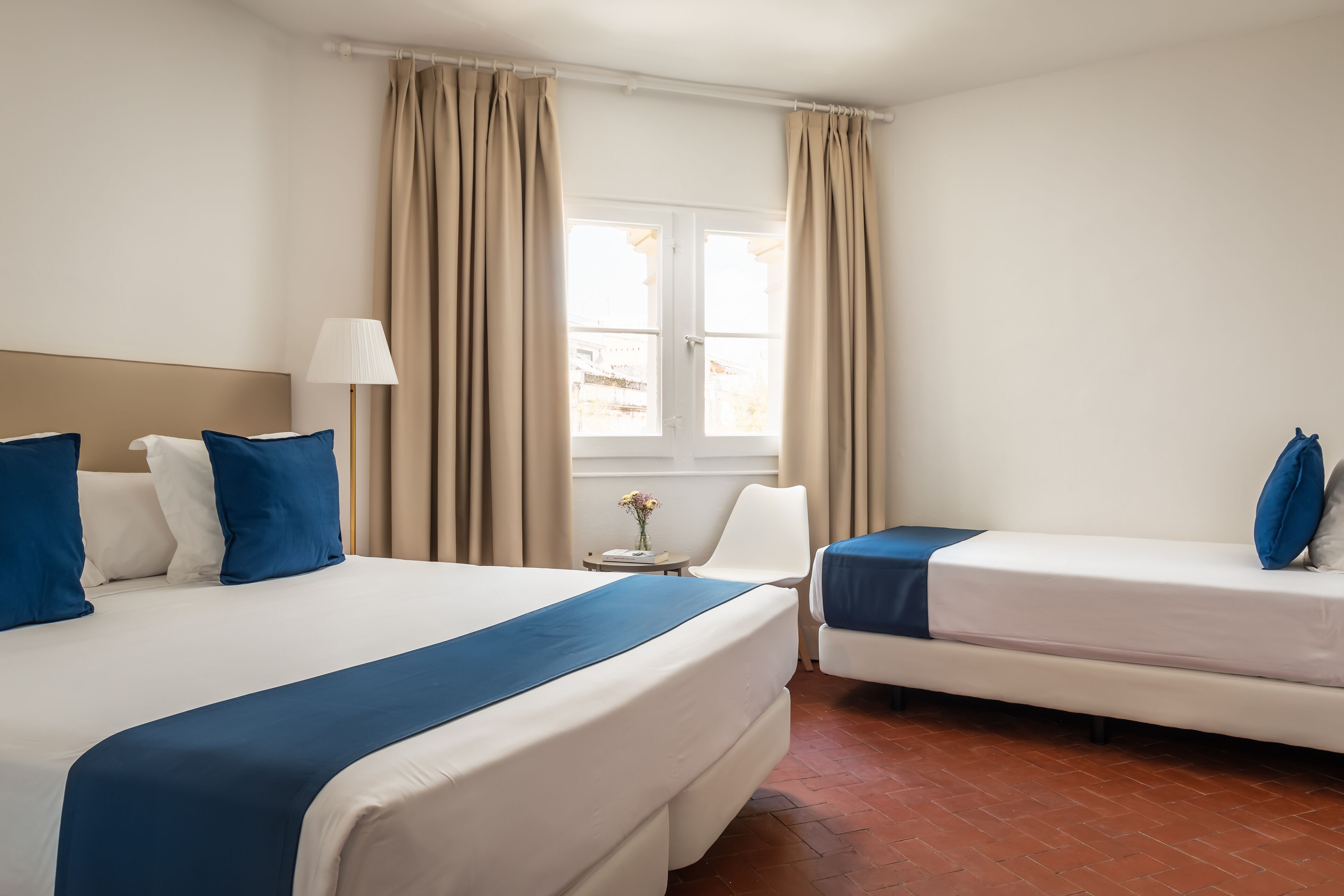 Ein Hotelzimmer mit zwei Betten, einem Fenster mit Vorhängen, einer Nachttischlampe und einem kleinen Tisch mit Blumen.
