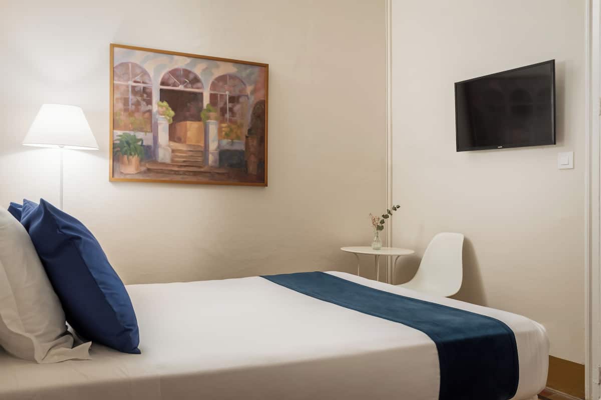 A hotel room with a bed, a painting, a TV, a lamp, and a small table with a vase.