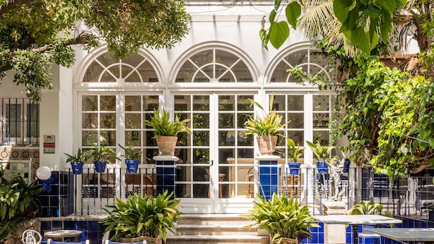 A courtyard with a white building featuring arched windows and a tiled floor. There are potted plants and outdoor seating areas.