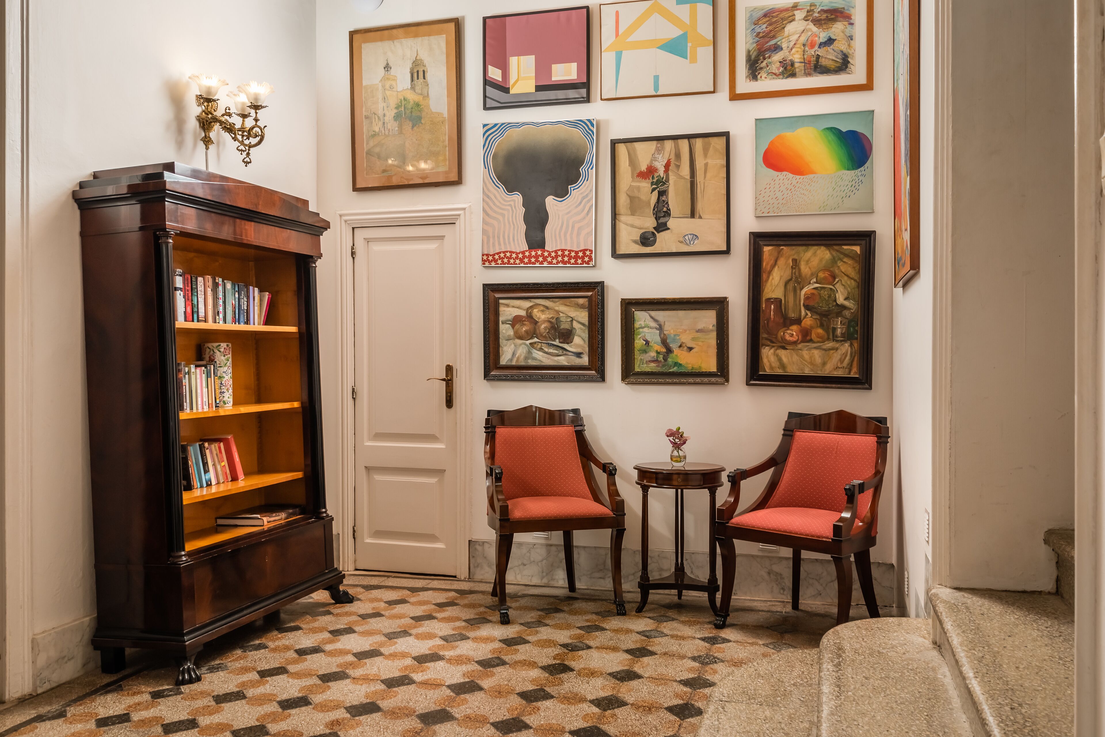 A room with a checkered floor, a bookshelf, two armchairs, and various framed artworks on the wall.