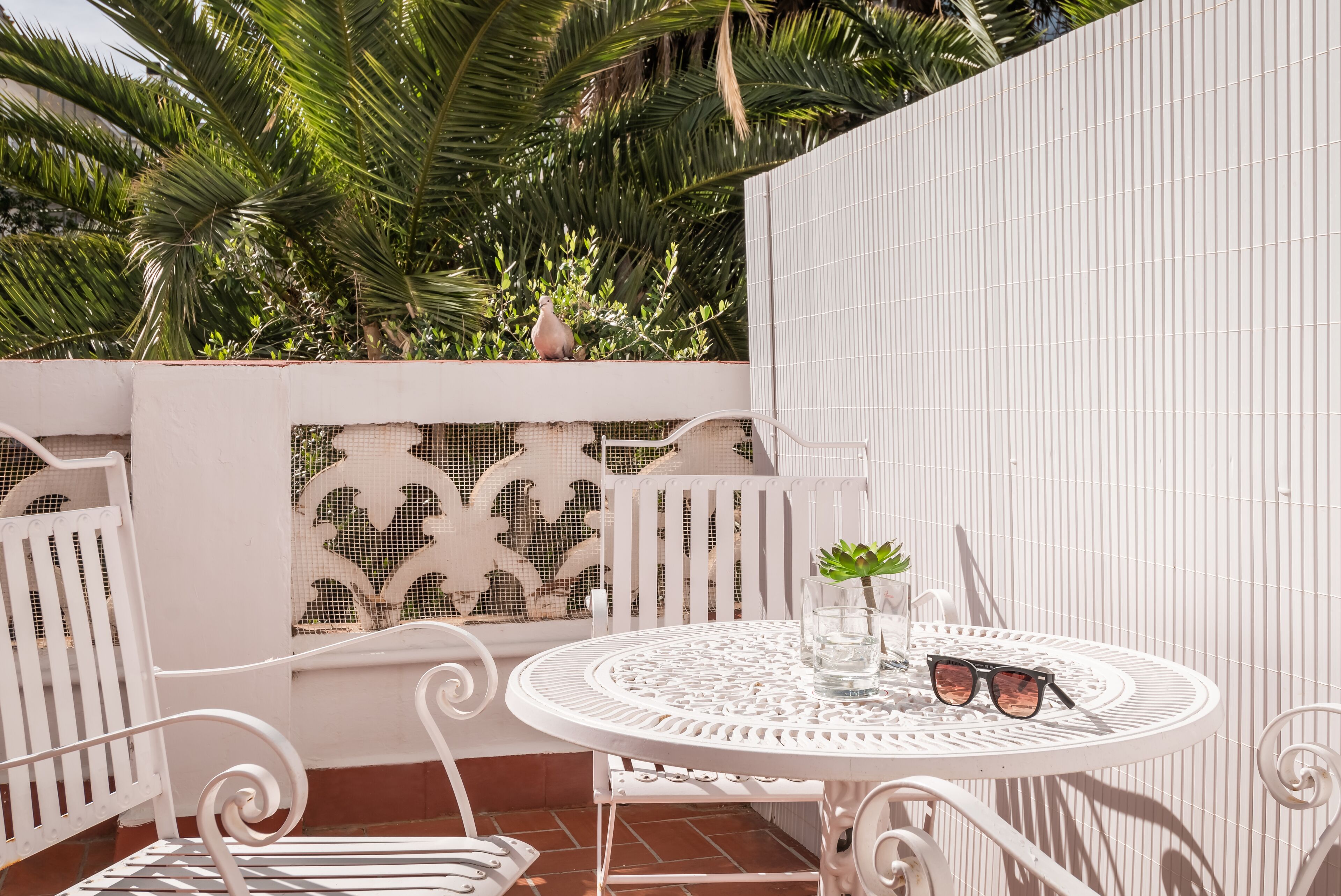 A round white metal table with two chairs, a small potted plant, and a pair of sunglasses on it.