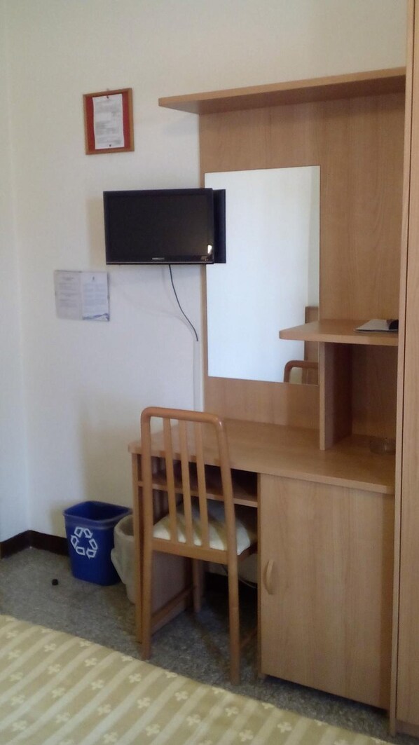 Um quarto pequeno com uma escrivaninha de madeira, uma cadeira, uma televisão fixada na parede e uma lixeira azul para reciclagem.