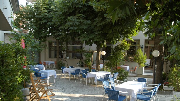 An outdoor café with tables and chairs arranged on stone pavement, surrounded by trees and buildings. - Moretti (Senigallia)