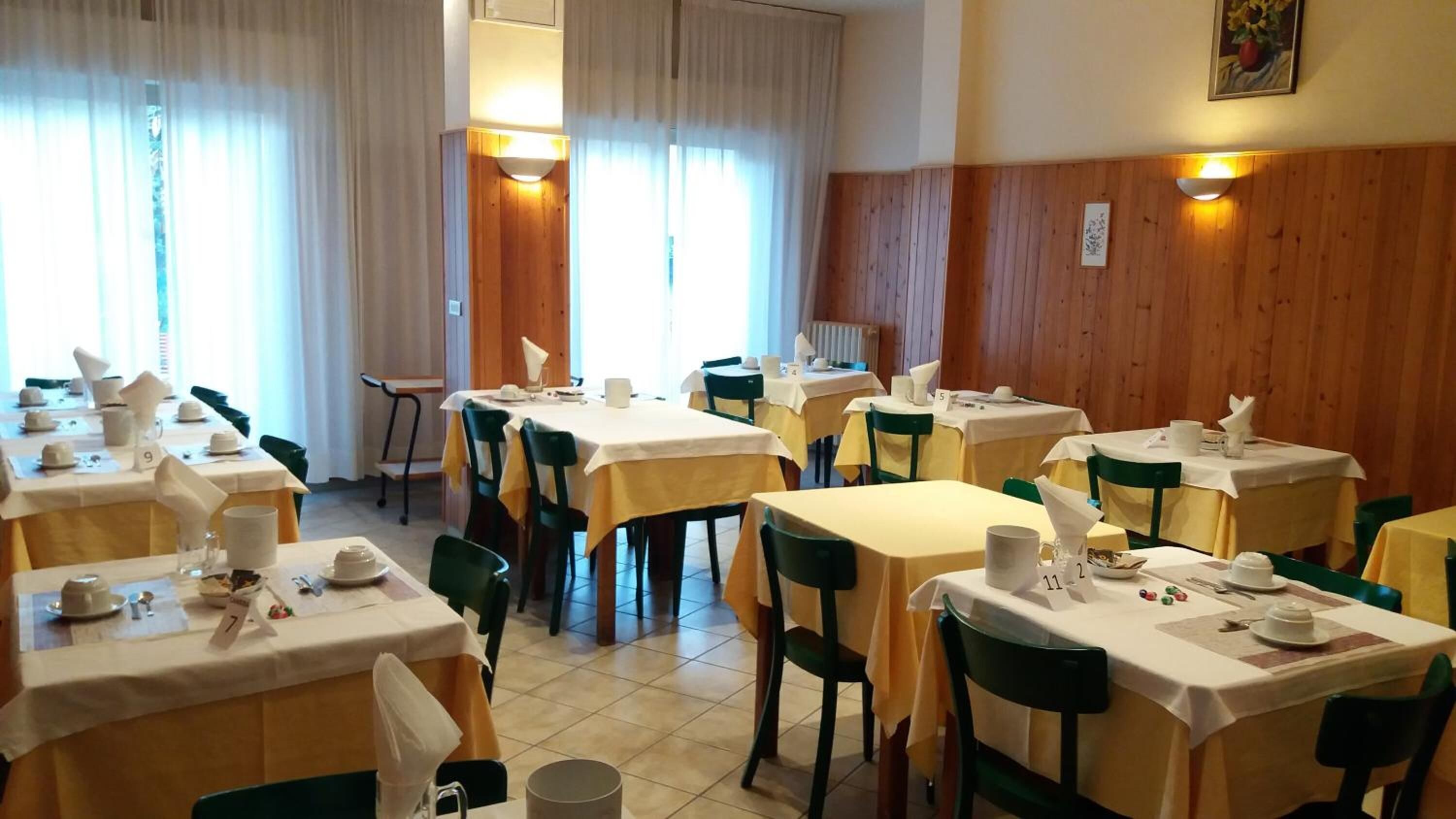 A dining area with multiple tables and chairs, arranged in rows. Each table is set with white tablecloths, plates, and cutlery. The room has wooden paneling on the walls and large windows with curtains.