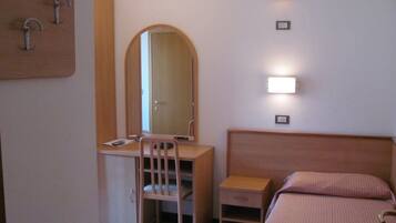 Una camera da letto con una scrivania in legno, una sedia e un letto con testiera. Si vede uno specchio sopra la scrivania e luci fissate al muro.