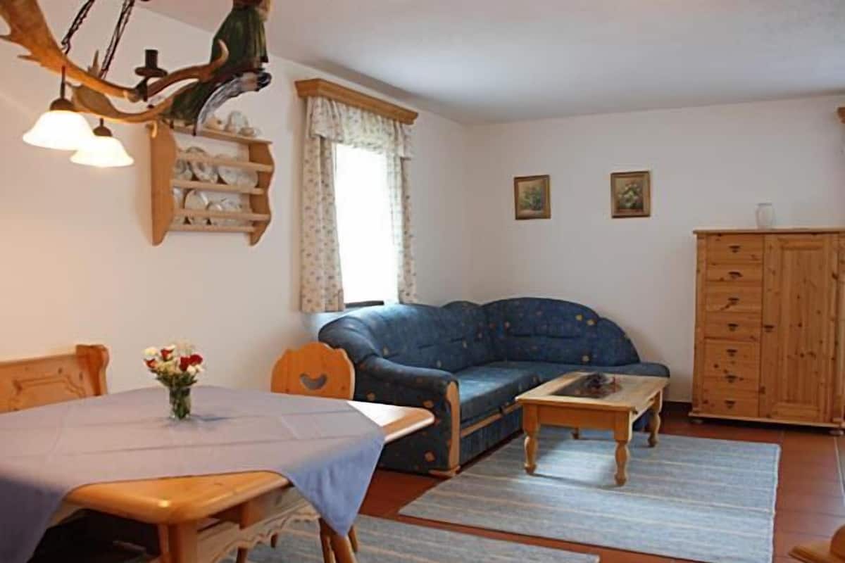 A cozy living room with a wooden dining table, a blue sofa, a wooden cabinet, and a small table with a vase of flowers.