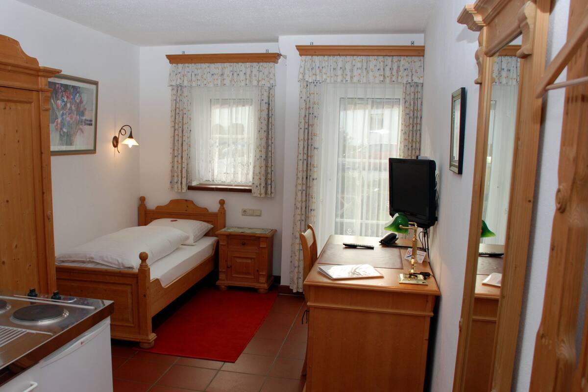 A compact bedroom with a wooden bed, a desk with a computer, a mirror, and a window with curtains.