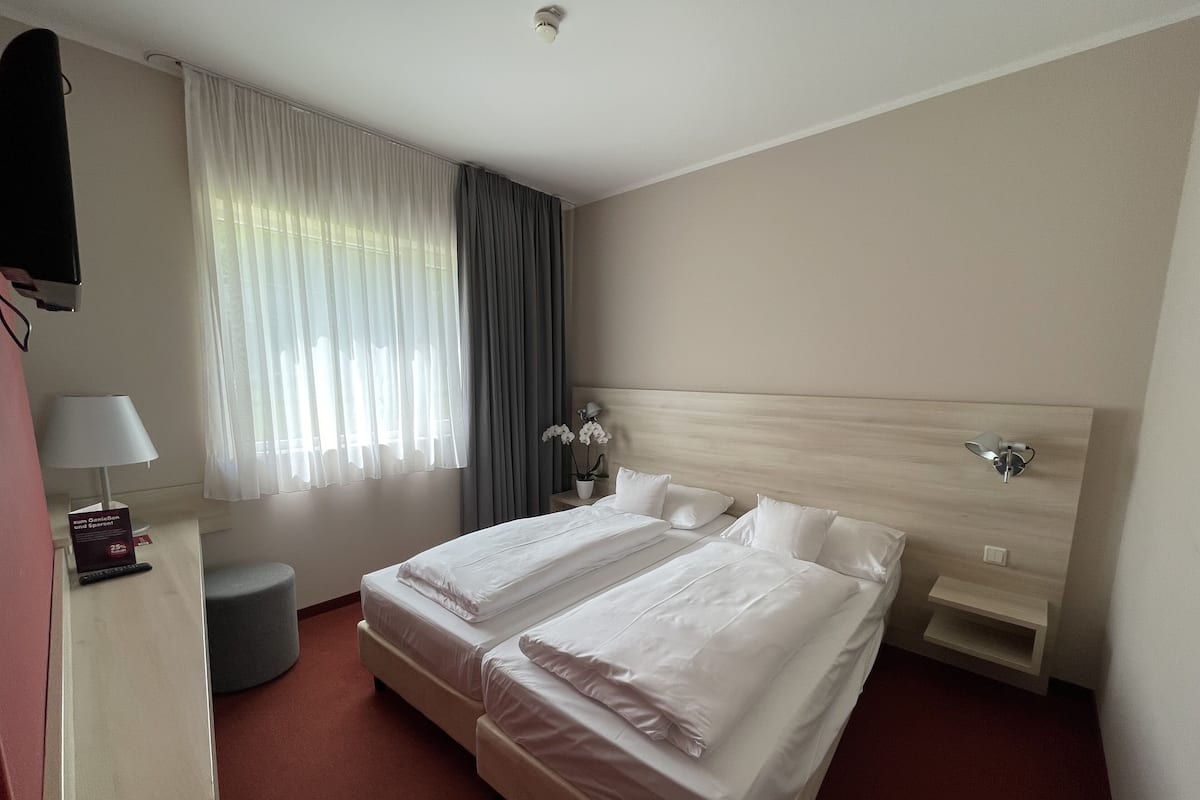 Ein Hotelzimmer mit zwei Betten, einem Nachttisch mit Lampe, einer Zimmerpflanze und einem großen Fenster mit Vorhängen.