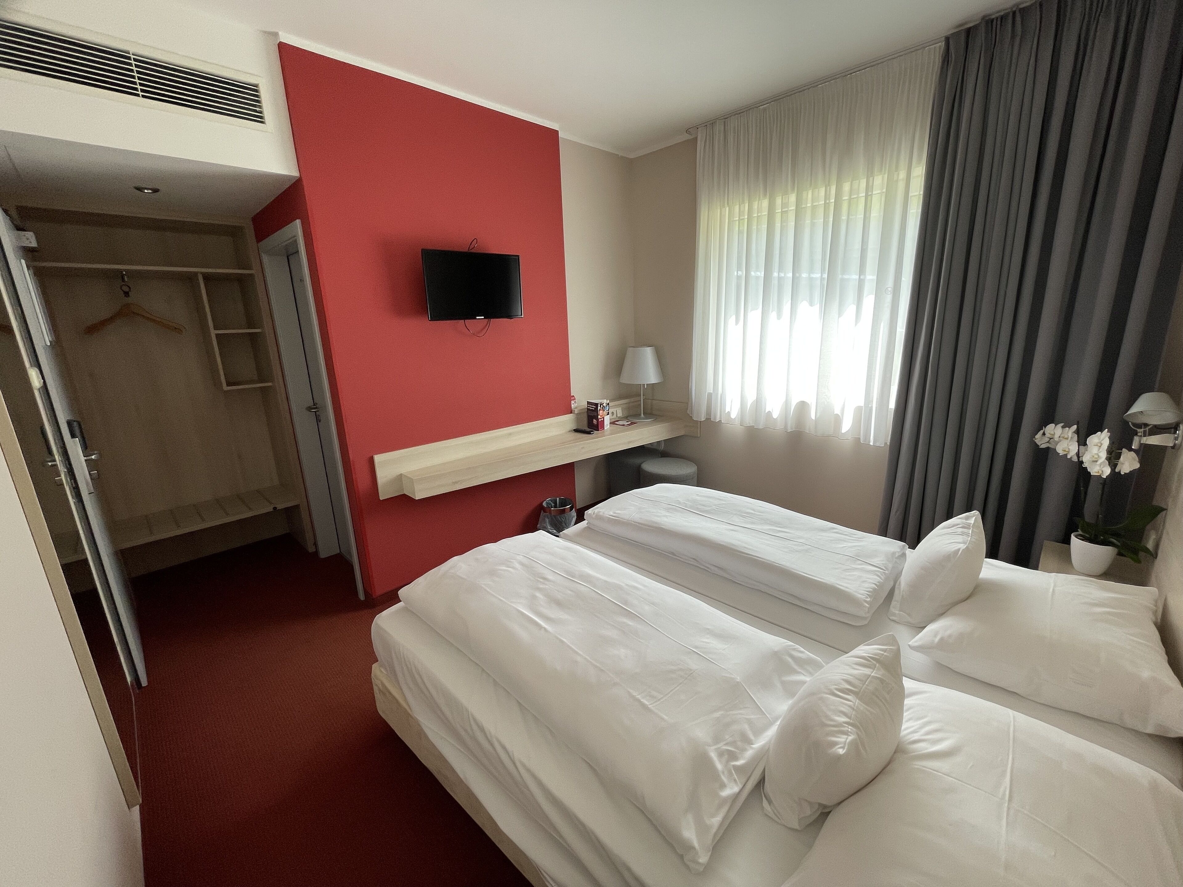 Camera d'albergo con due letti, una televisione fissata al muro, un comodino con una lampada e una finestra con tende.