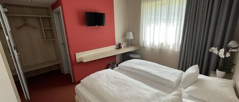 A hotel room with two beds, a TV mounted on the wall, a bedside table with a lamp, and a window with curtains.