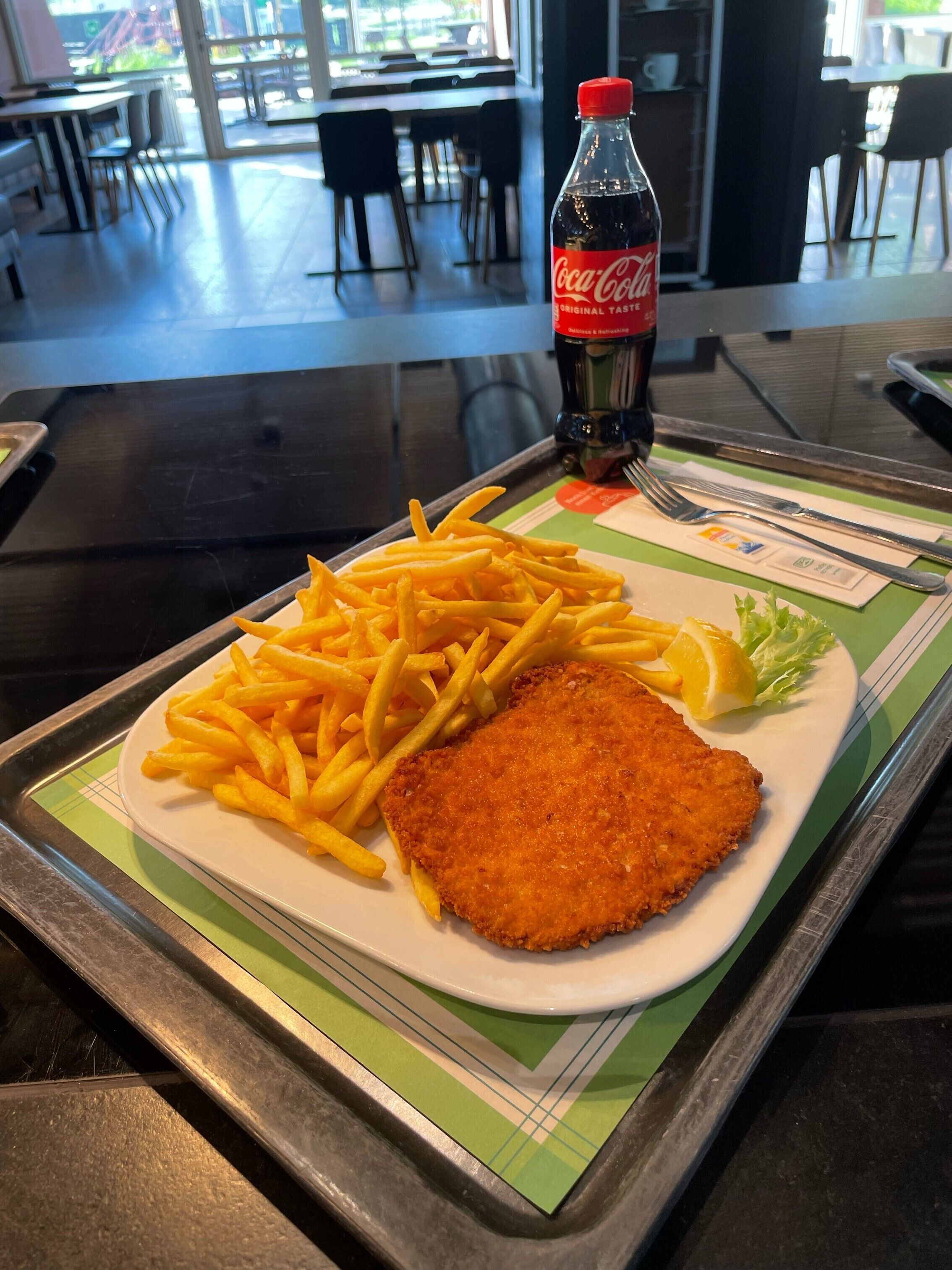 A meal consisting of a breaded cutlet, French fries, and a lemon wedge on a white plate, with a Coca-Cola bottle in the background.