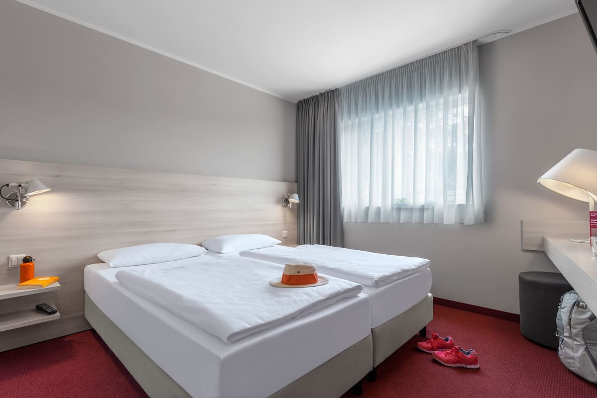 Ein Hotelzimmer mit zwei Betten, einem Schreibtisch, einem Stuhl, einer Lampe, einem Koffer und einem Fenster mit Vorhängen.