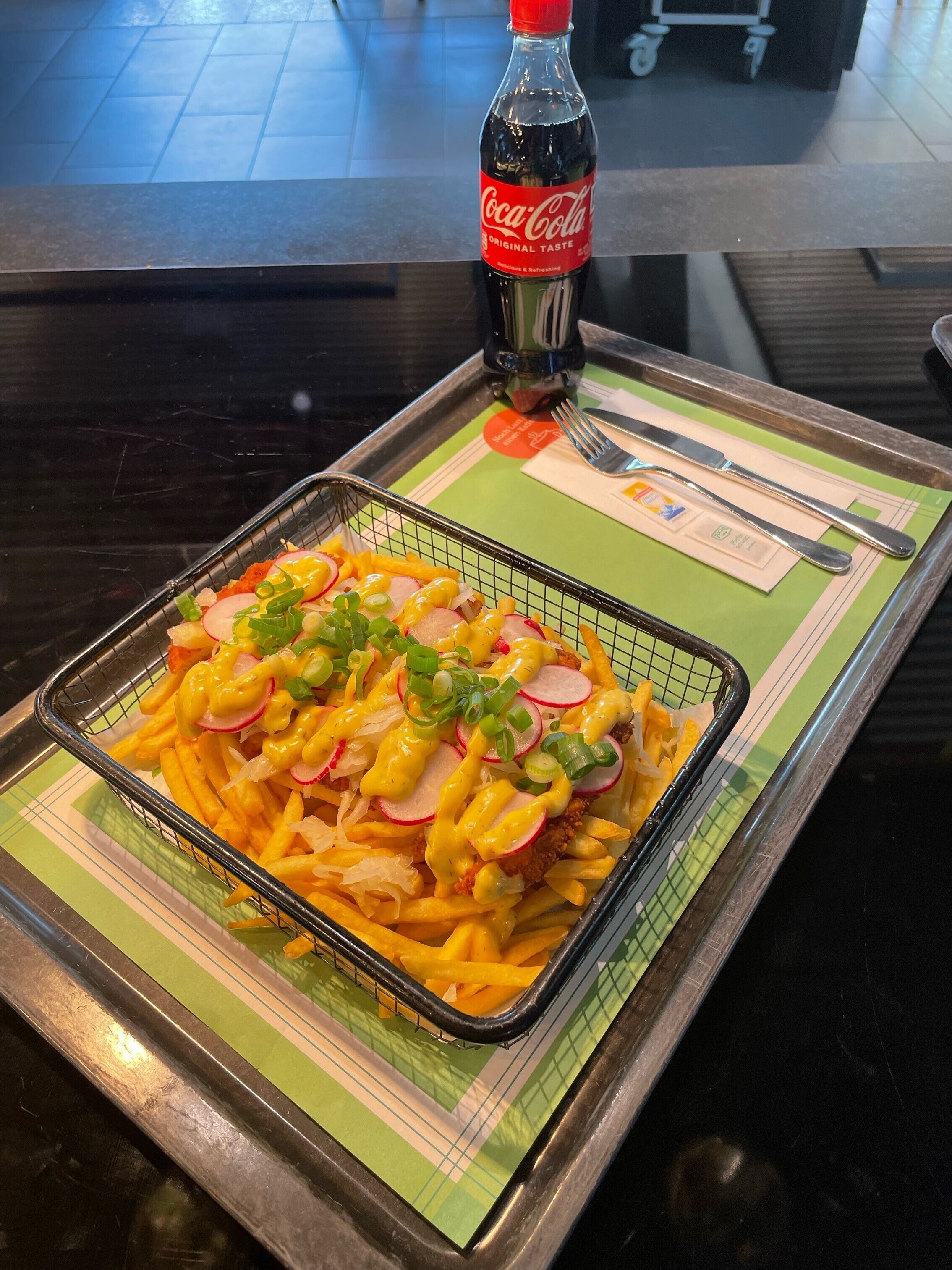 A tray of nachos with cheese, onions, and green onions, a bottle of Coca-Cola, and a fork and knife on a green placemat.