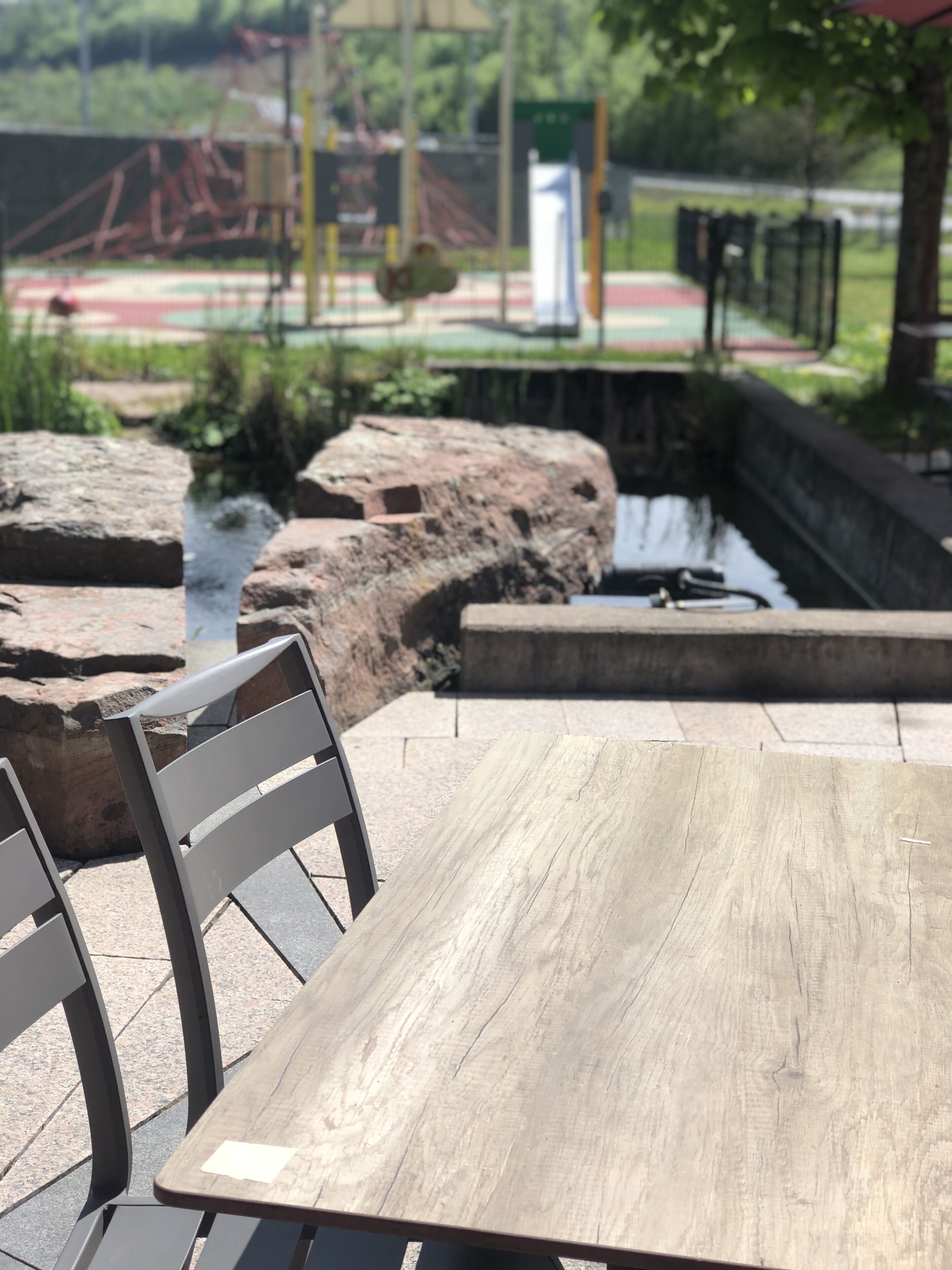 Mesa de madera con sillas de metal, ubicada al aire libre con vista a un parque y un puente al fondo.