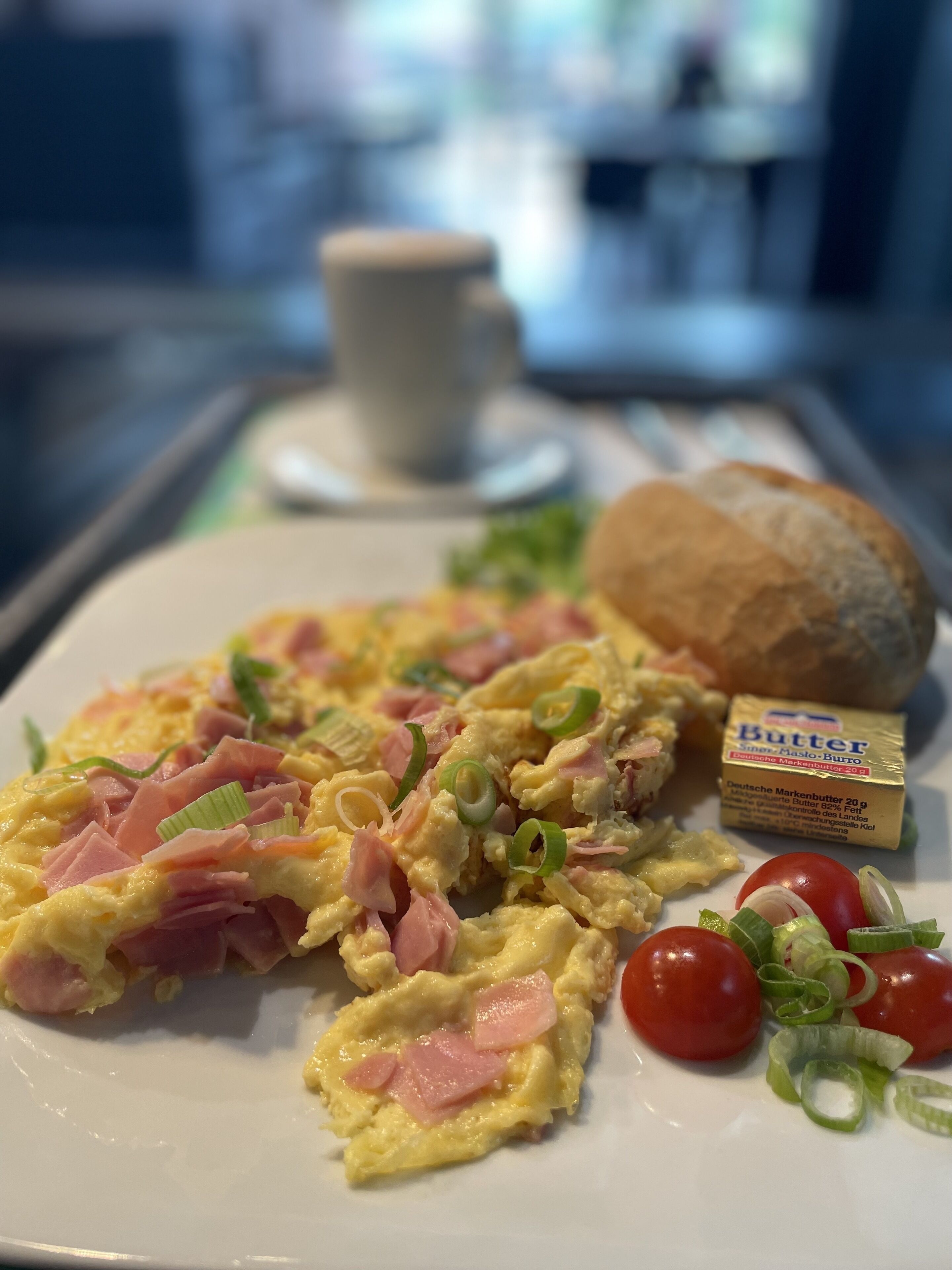 A breakfast plate with scrambled eggs, ham, and green onions, accompanied by a slice of butter, a small bread roll, and cherry tomatoes.