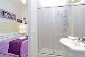 Un dormitorio con cama, mesita de noche con lámpara y baño con ducha acristalada y lavamanos.