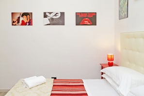 Una habitación de hotel con una cama, una colcha de rayas rojas, un almohadón blanco y una mesita de noche con una lámpara roja. Hay tres cuadros en la pared.