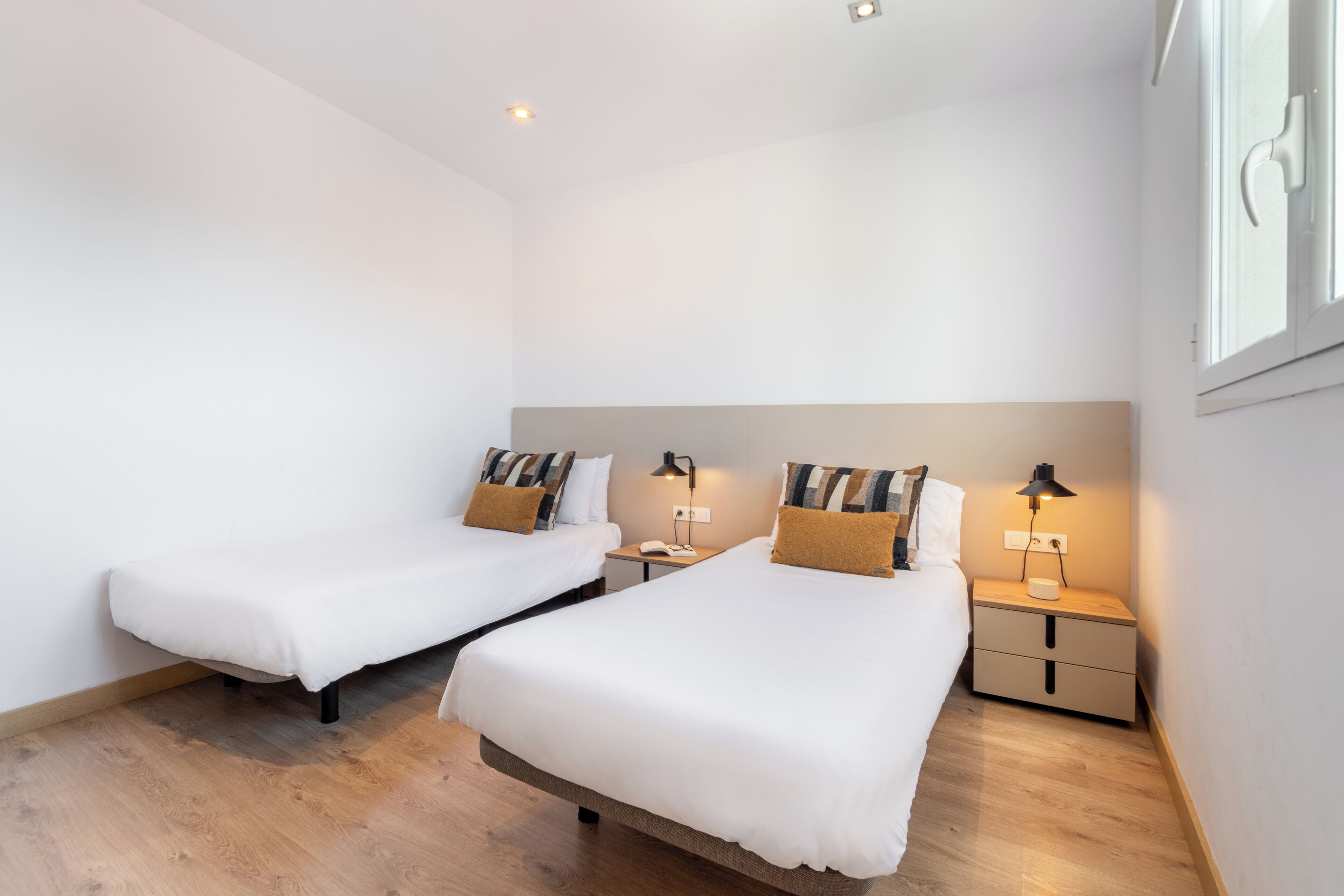 A room with two single beds, bedside tables with lamps, a window, and a wooden floor.
