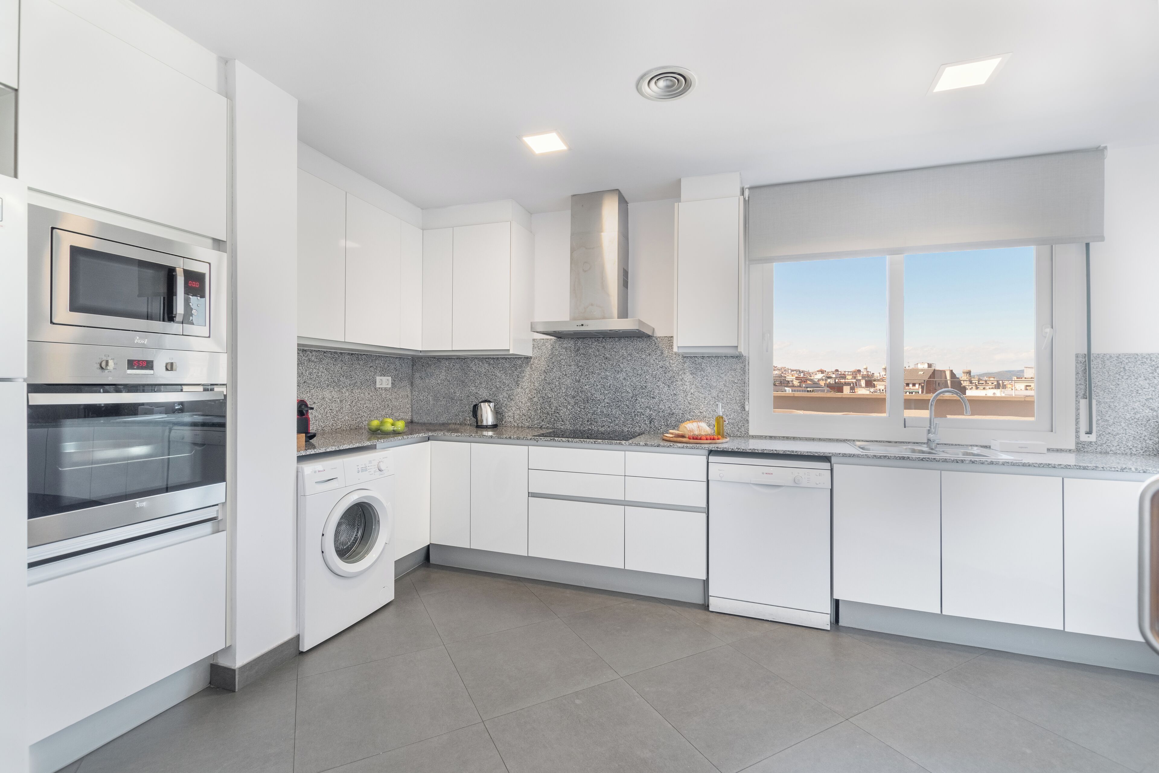 Una cocina moderna con armarios blancos, horno empotrado, microondas y lavavajillas, encimera de granito y ventana con vista a la ciudad.