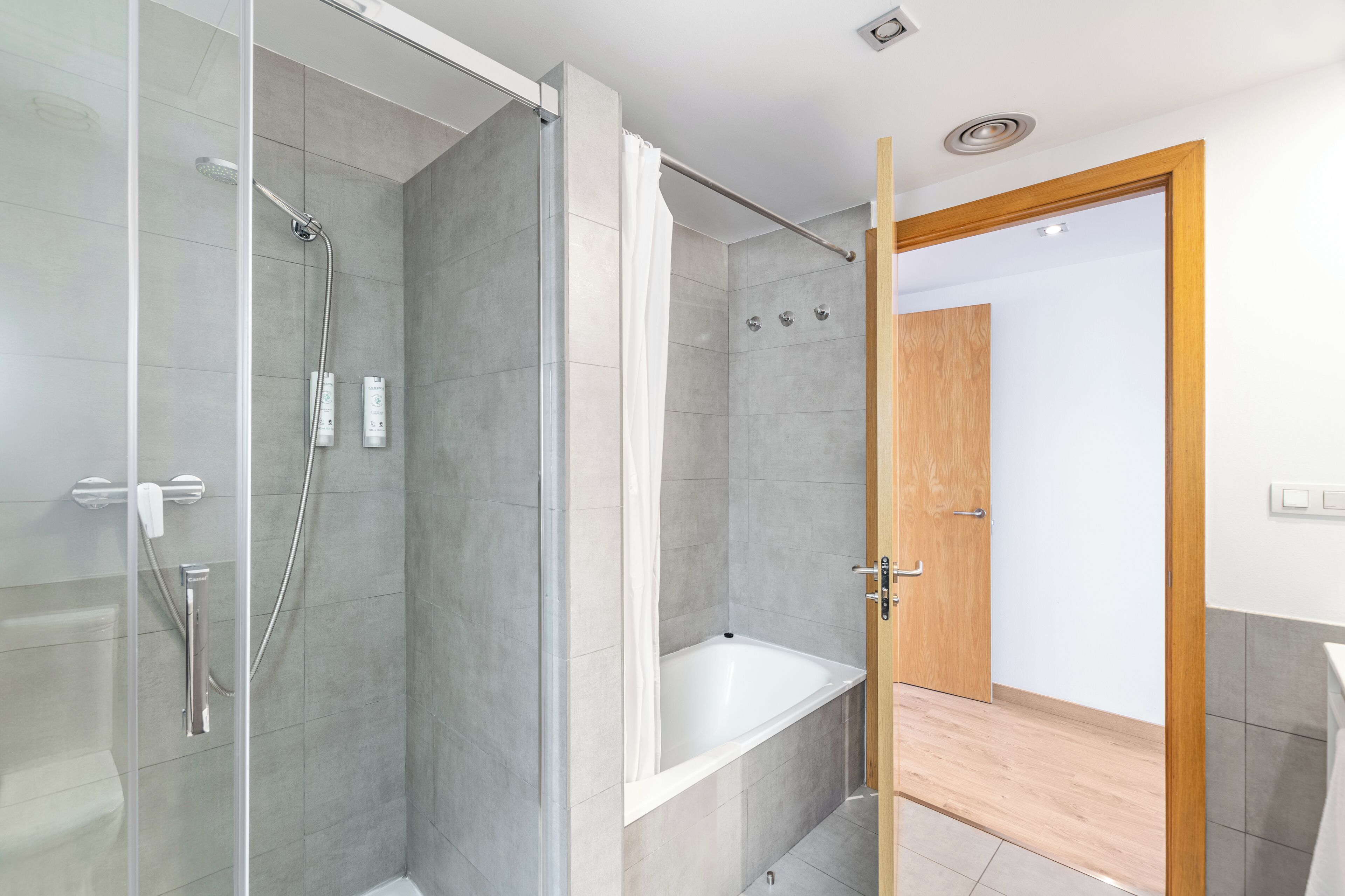 A modern bathroom with a glass-enclosed shower, a white bathtub, and a wooden door leading to another room.