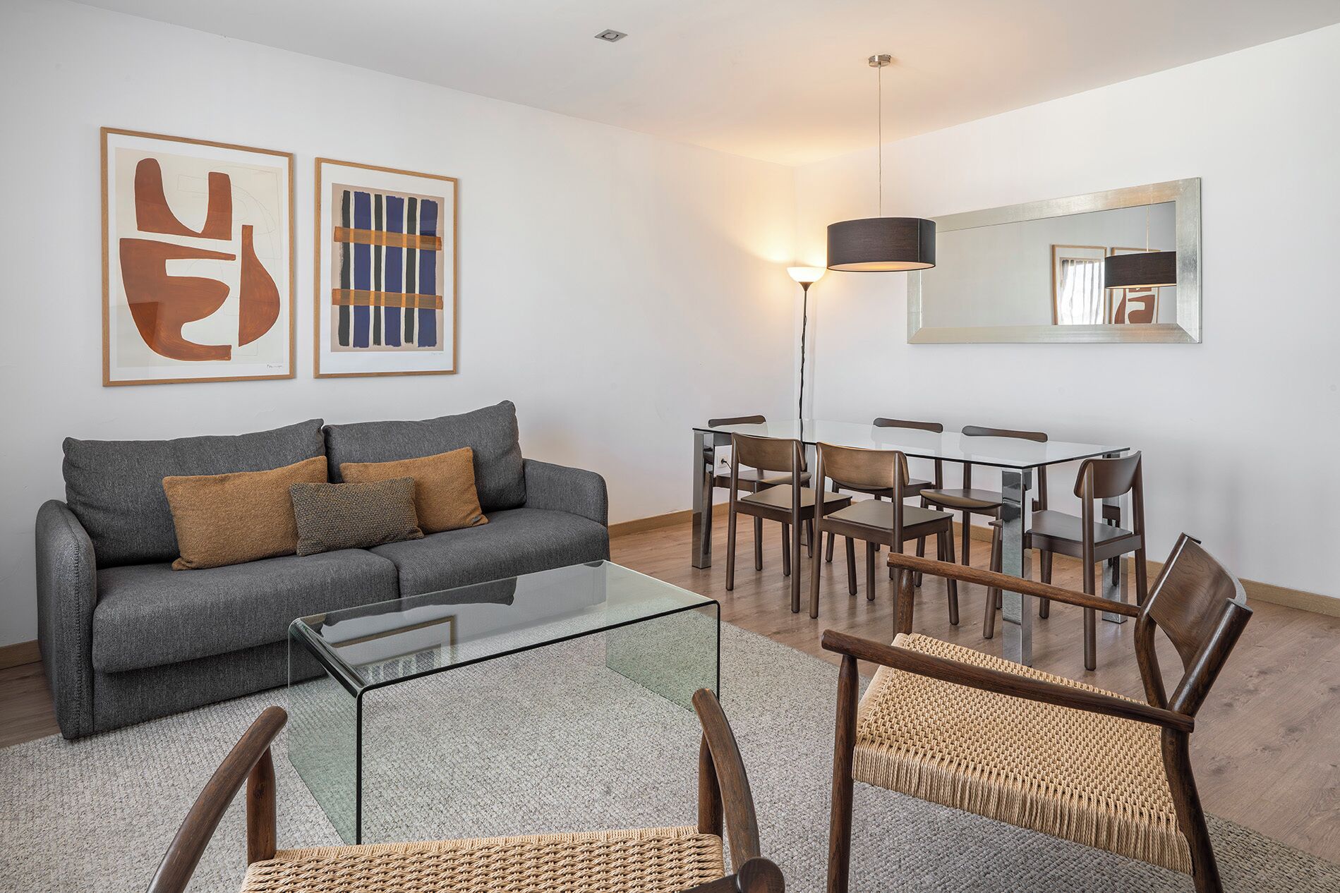 A living room with a grey sofa, a glass coffee table, wicker chairs, a dining table with chairs, and abstract art on the wall.