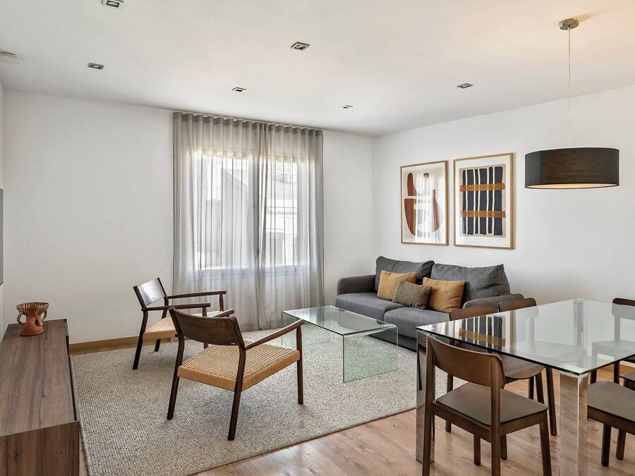 Ein modernes Wohnzimmer mit einem Glastisch, Holzstühlen, einem grauen Sofa mit Kissen, einem großen Fenster mit transparenten Vorhängen und abstrakter Kunst an der Wand.