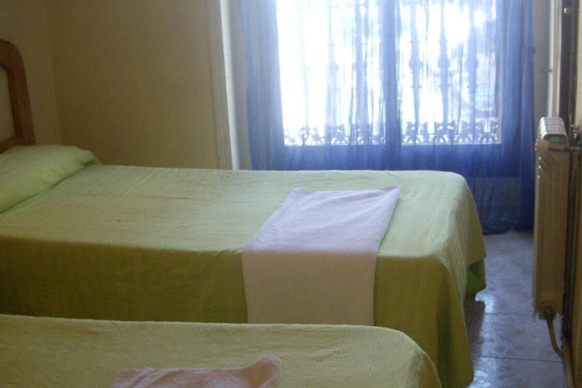 A room with two single beds, each with a green bedspread and a white pillow. There is a window with blue curtains, and a radiator is visible on the right side.