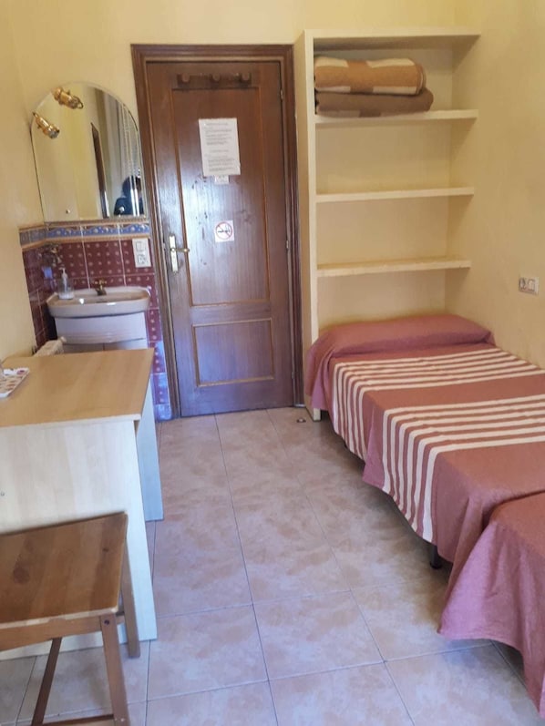 A small, simple room with a wooden desk, a sink, a mirror, a door with a notice, and two beds with striped bedding. - Hostal Pacios (Madrid)