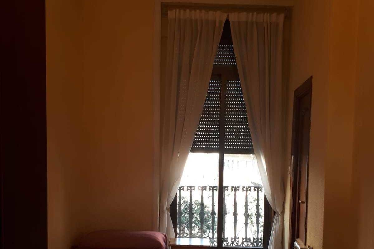 A room with two beds, a wooden table, and a window with a view of a balcony.