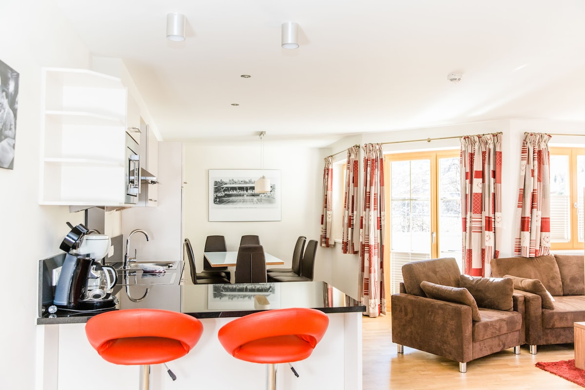 A modern kitchen with a coffee machine, dining table, and chairs. There is a living area with a sofa and red chairs.