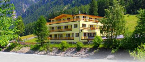 A multi-story building with balconies, located near a river with a mountainous backdrop.