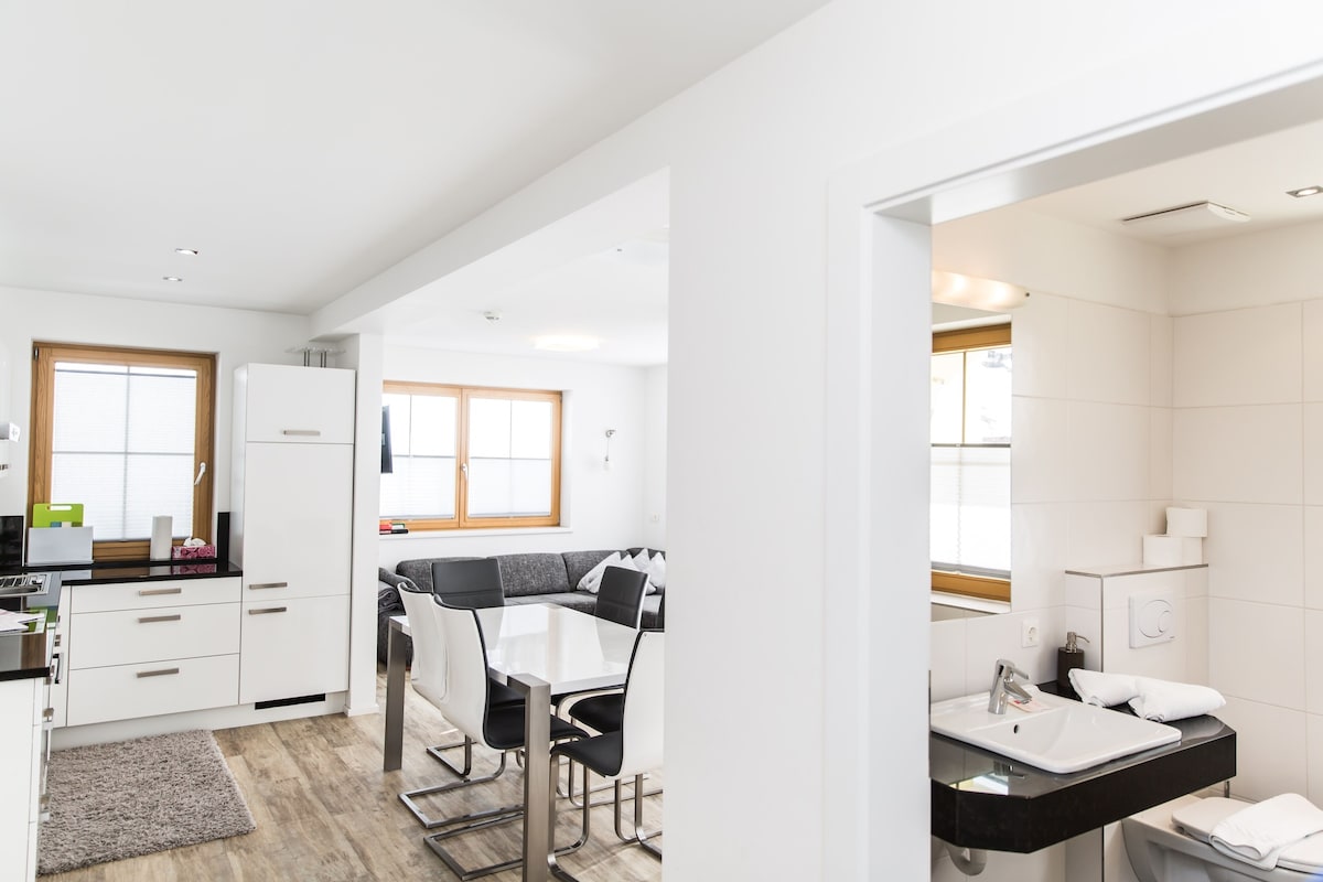 A modern kitchen with white cabinets, a dining area with a black and white table, and a bathroom with a white sink and mirror.