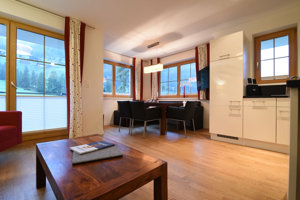 A modern kitchen with a dining area, wooden flooring, and large windows offering a view of the outdoors.