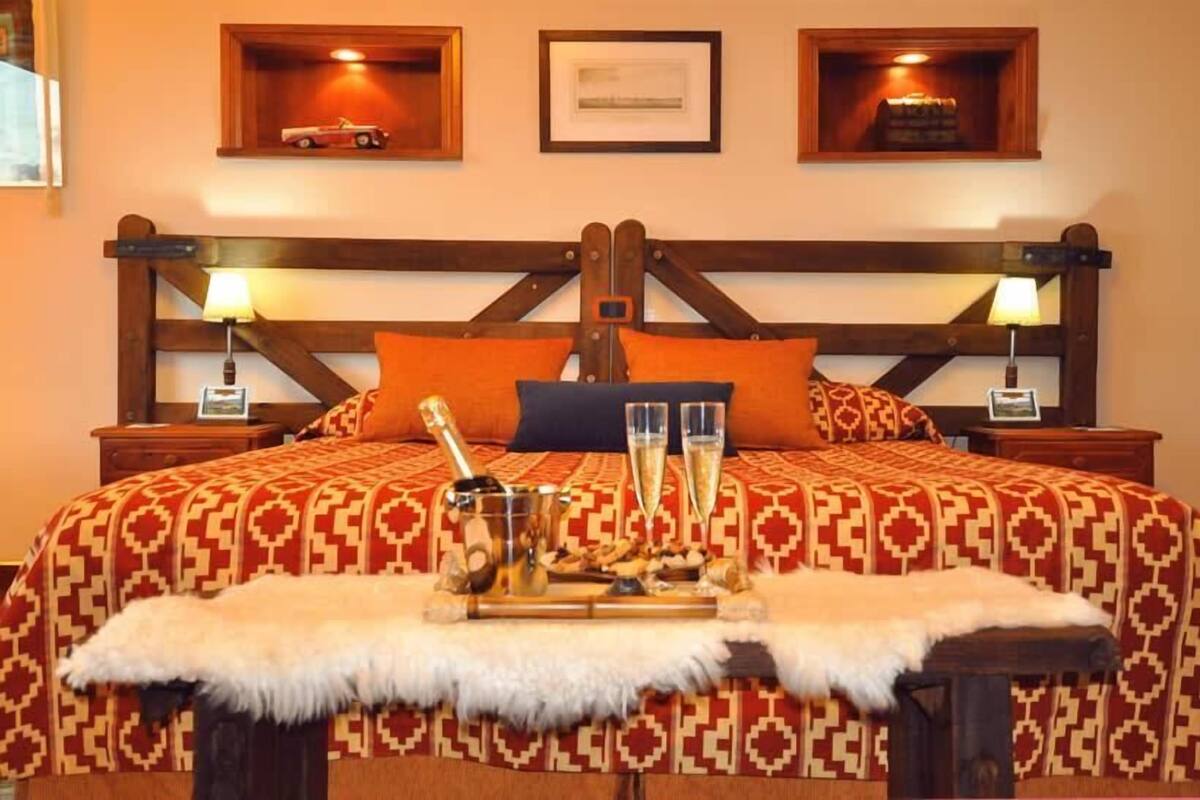 A bed with a patterned bedspread, two pillows, a tray with glasses and a bottle, and a fur throw.
