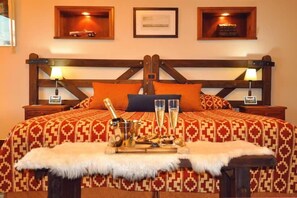 Una cama con una colcha estampada, dos almohadas, una bandeja con vasos y una botella, y un plaid de piel.