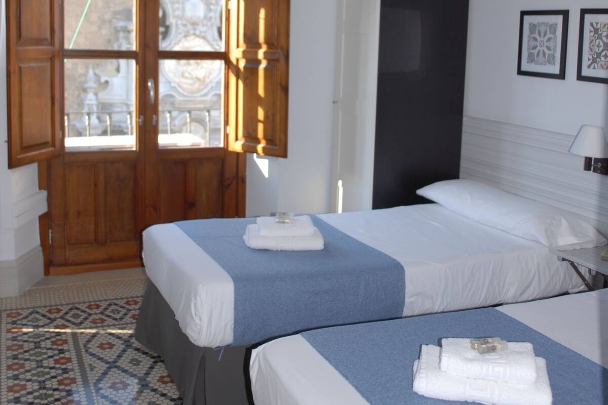 Quarto de hotel com duas camas, piso estampado, uma janela grande com persianas de madeira e um lustre.