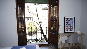Habitación de hotel con cama, escritorio, silla, balcón con vistas a edificios y árboles, y un cuadro en la pared.