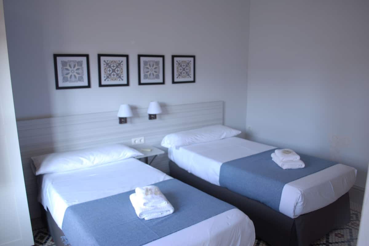 Quarto de hotel com duas camas, cada uma com colcha azul e lençóis brancos. Há quadros nas paredes acima das camas e uma mesinha com duas luminárias entre elas.