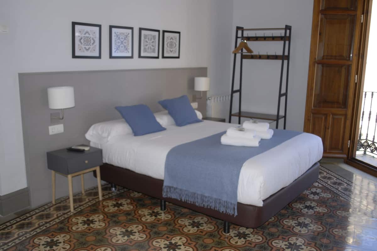 Cama bem arrumada com almofadas azuis, mesa de cabeceira com abajur e um cabideiro.