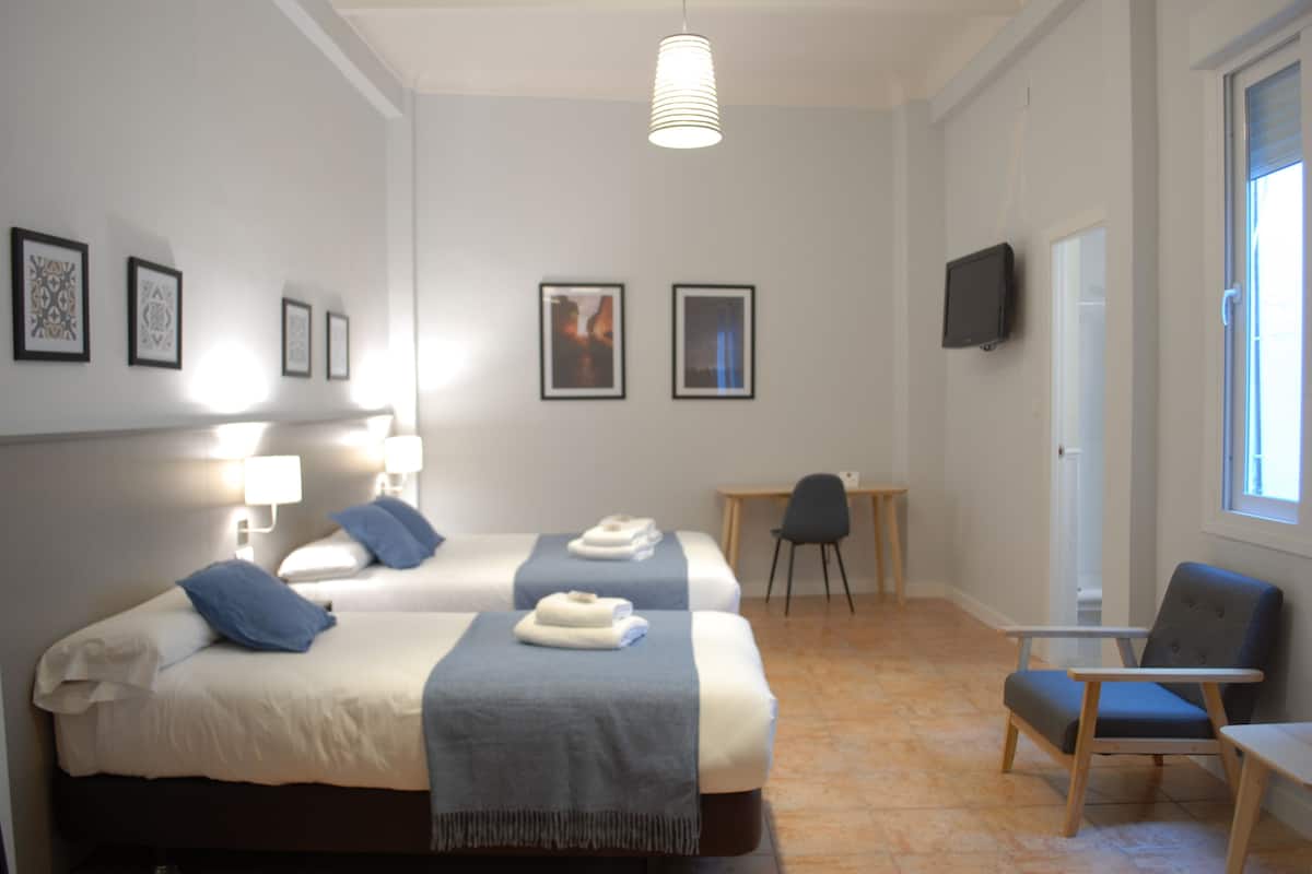 Quarto de hotel com duas camas, uma escrivaninha, uma cadeira, uma televisão e quadros na parede.