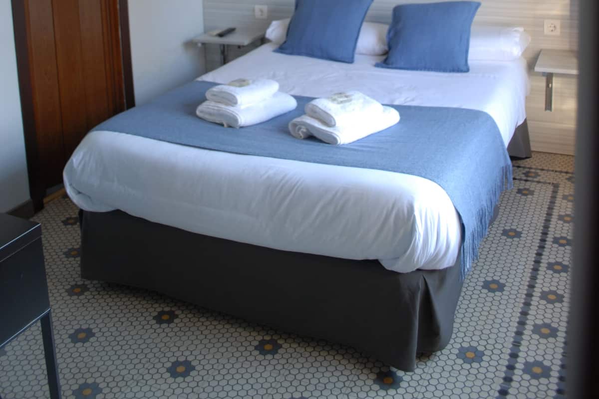Quarto de hotel com uma cama de casal, duas mesas de cabeceira, um criado-mudo, uma porta de madeira e piso ladrilhado.
