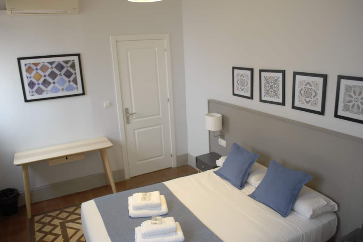Quarto de hotel com uma cama, escrivaninha, cadeira, mesa de cabeceira, abajur e porta.