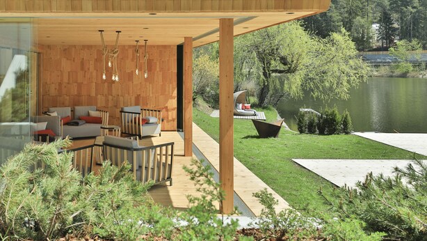 Un área de estar moderna al aire libre con suelo de madera, pared de cristal y vista a un lago.