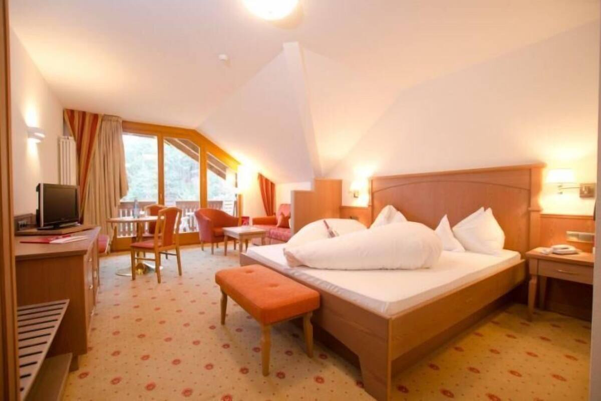 Ein Hotelzimmer mit einem großen Bett, einem Schreibtisch mit Computer, einem Stuhl, einem kleinen Tisch und einem Fenster mit Vorhängen.