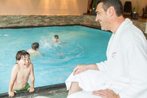 A pool area with a person in a white robe sitting by the pool, a child in swim trunks, and another person swimming. - Hotel Weihrerhof (Renon)