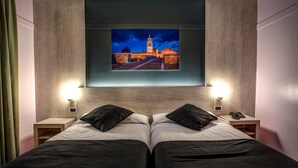 Une chambre d’hôtel avec deux lits, un grand écran de télévision au-dessus des lits diffusant une vue sur la ville, des lampes de chevet et une tête de lit en bois.