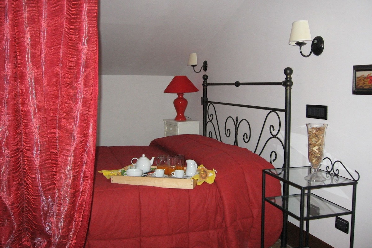 Un dormitorio con una colcha roja, una mesita de noche con una lámpara roja, una mesa de centro de cristal y una bandeja con objetos para té.