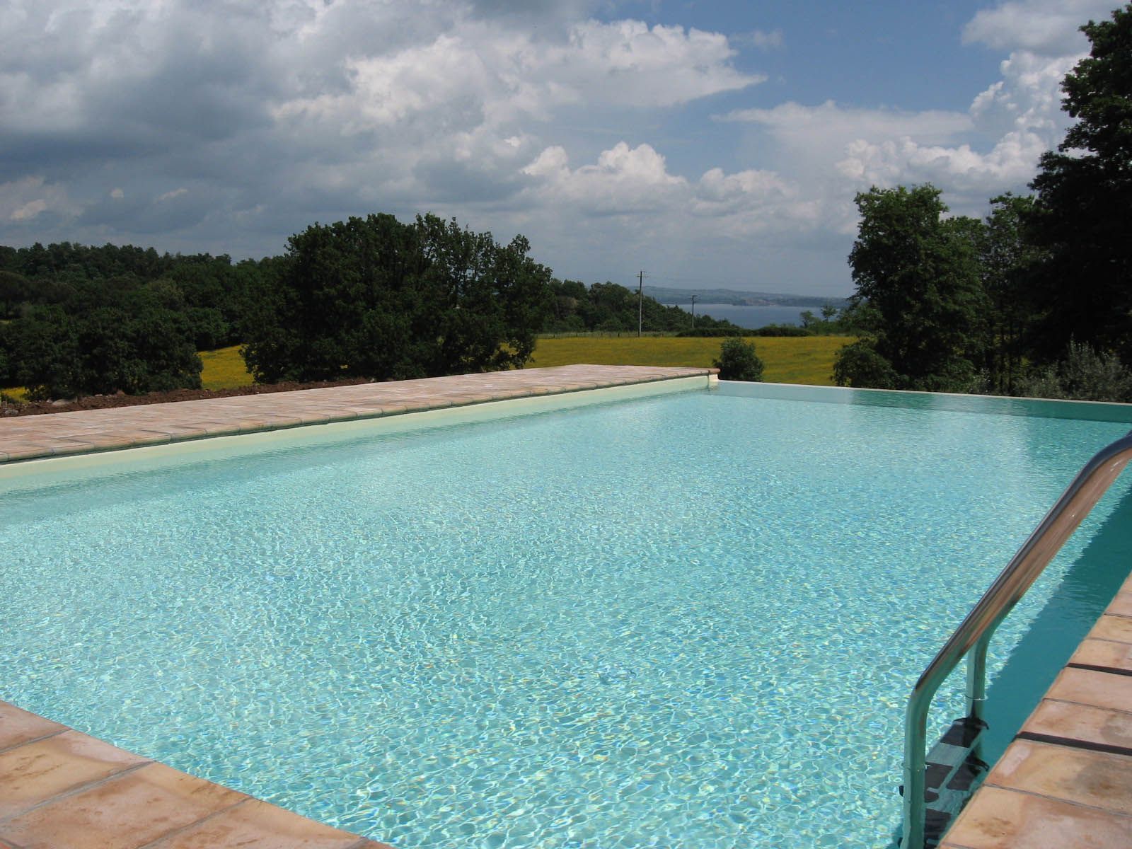 A rectangular swimming pool with clear blue water, surrounded by a paved deck and lush greenery.
