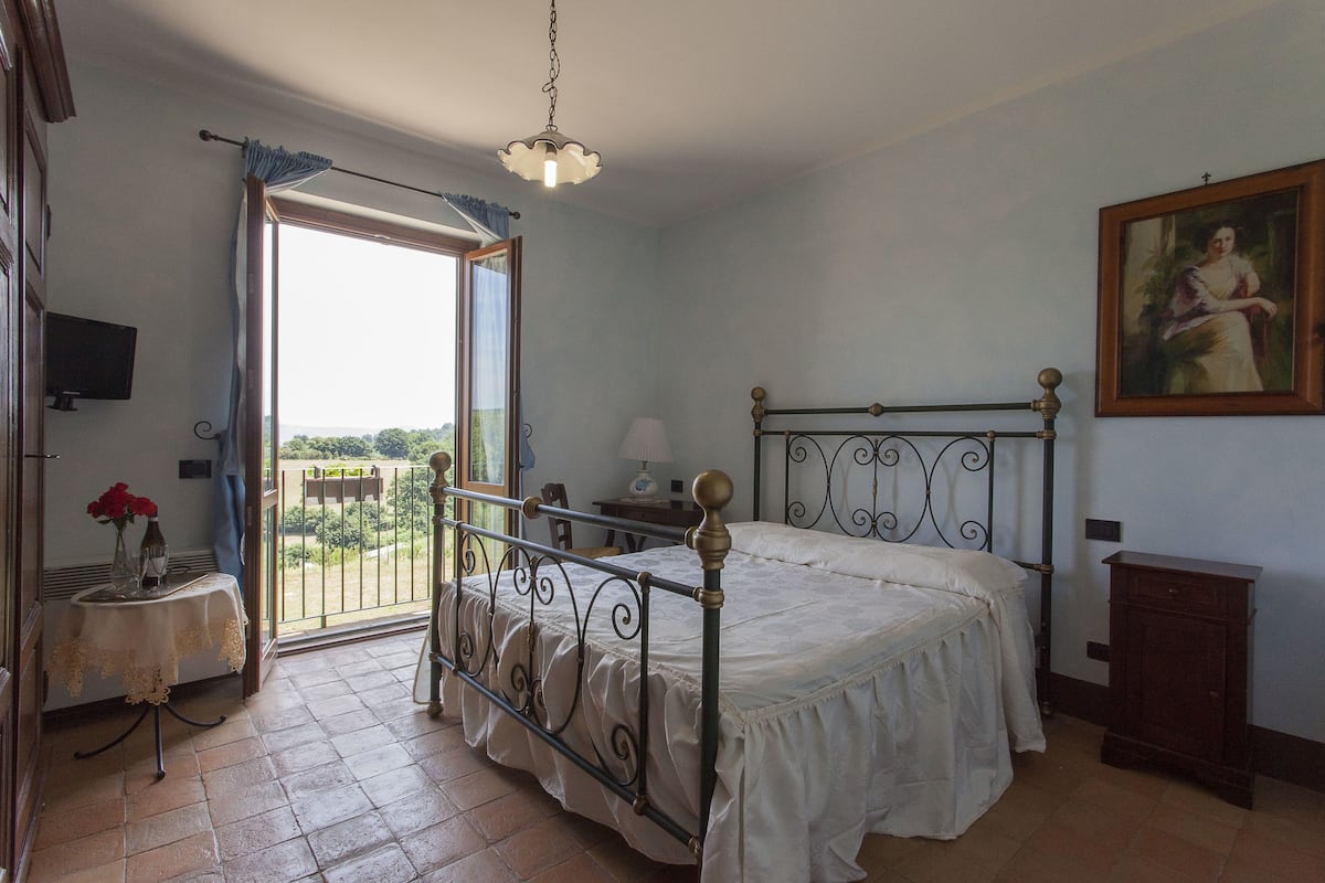 Un dormitorio con cama, una mesa con flores, una lámpara y un cuadro en la pared.